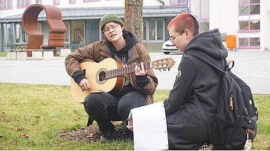 Eine Person hockt vor einem Baum und spielt Gitarre, eine zweite hält einen Block