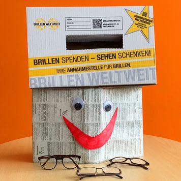 Brillensammelbox in gelb-weiß auf einem Buch mit Smiley
