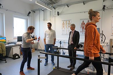 Frau läuft auf Laufband, drei Personen unterhalten sich im Hintergrund