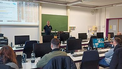 Teilnehmende vor Ort und Online folgen dem Vortrag