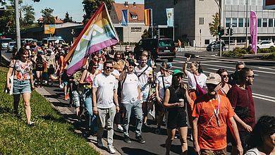 Viele Menschen laufen am Rand einer Straße, jemand schwenkt eine große Regenbogenfahne