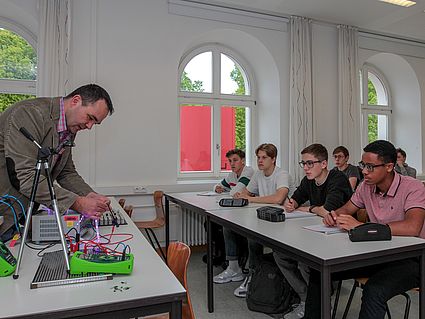 Schüler sitzen im Hörsaal, Professor zeigt ihnen den Aufbau einer Transistorensteuerung 