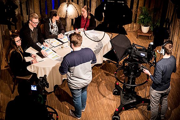 Studenten stehen um einen Tisch herum und arbeiten im TV-Studio. Ein Kameramann nimmt auf.