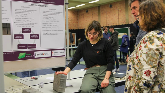 Junge Frau in schwarzem Shirt mit Logo der OTH AW präsentiert anhand eines Betonklotzes ihr Projekt zur Speicherung von Co2. Im Hintergrund sieht man eine Stellwand, auf der Informationen zum Projekt dargesteltt sind (mit Logo von Jugend Forscht und der Firma Godelmann).
