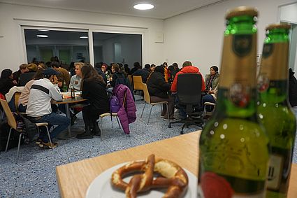 Im Vordergrund sieht man einige Flaschen Bier und eine Breze, im Hintergrund sitzen um die 20 Menschen an kleineren 