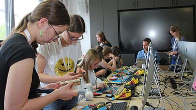 Mädchen sitzen an einem Tisch mit Bildschirmen, Lötkolben und anderem Werkzeug