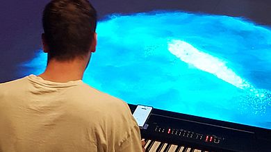 Mann von hinten sitzt am Keyboard und vor ihm ist eine blaue Fläche auf dem Boden projiziert.