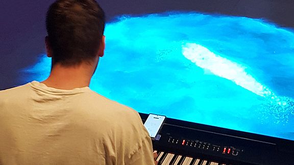 Mann von hinten sitzt am Keyboard und vor ihm ist eine blaue Fläche auf dem Boden projiziert.