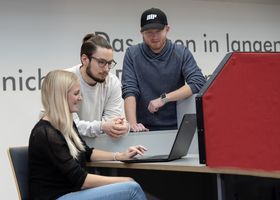 [Translate to Englisch:] Drei Studierende schauen in der Bibliothek auf den Bildschirm eines Laptops