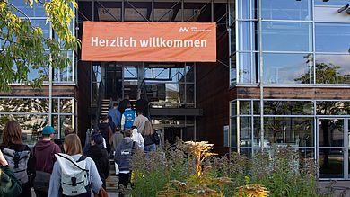 Junge Menschen gehen in ein Gebäude, über der Eingangstür hängt ein großer "Herzlich willkommen"-Banner