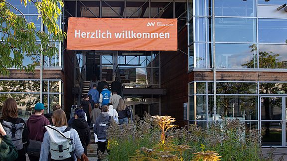 Junge Menschen gehen in ein Gebäude, über der Eingangstür hängt ein großer "Herzlich willkommen"-Banner