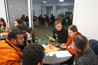 sechs Personen sitzen um einen Tisch und reden, im Hintergrund sind weitere Studierende