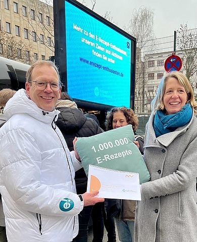 Ein Mann reicht einer Frau ein Kissen mit der Aufschrift "1.000.000 E-Rezepte" und eine gebundene Studie.  