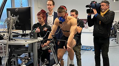 Ein Mann fährt auf einem Ergometer, mehrere Personen blicken auf einen Bildschirm. Ein weiterer Mann filmt.