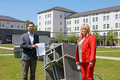 Andrea Klug und Raphael Lechner stehen am Campus mit Berufungsurkunde