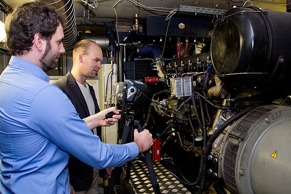 Am Blockheizkraftwerk-Prüfstand können vollständige BHKW-Systeme mit verschiedenen Kraftstoffen getestet werden. Zwei Laboringeniere stehen vor dem Blockheizkraftwerk-Prüfstand.