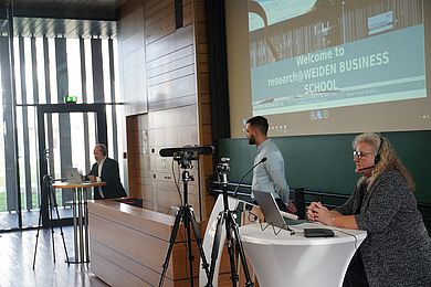 Drei Personen mit Laptops stehen in einem Hörsaal 